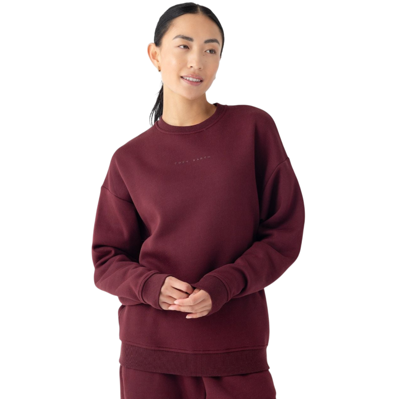 A person with dark hair tied back is wearing the Cozy Earth Women's CityScape Crewneck, paired with matching pants. They are standing against a plain white background. The crewneck features subtle lettering on the front. The person is smiling gently and has their hands clasped together. |Color:Burgundy