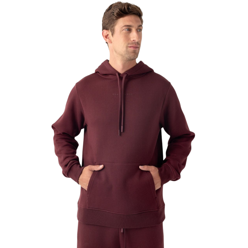 A man is wearing a maroon Men's CityScape Hoodie by Cozy Earth, looking slightly to the left. He has short brown hair and his hands are tucked into the front pocket of the hoodie. The background is plain white. |Color:Burgundy