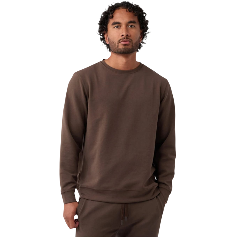 A man with curly hair and a beard wears the Men's StretchTech Crewneck by Cozy Earth in brown, along with matching pants. He stands against a plain white background with his hands in his pockets, looking straight at the camera. |Color:Cacao