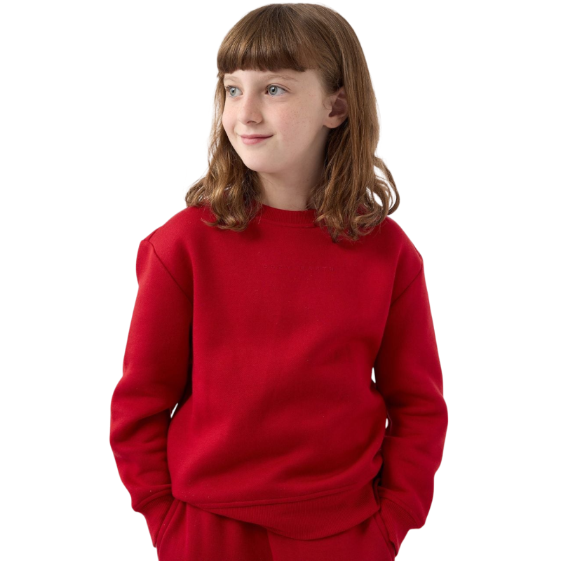 A young person with brown hair stands wearing a bright red Kid's CityScape Crewneck from Cozy Earth and pants against a plain white background, looking to the side and smiling slightly, with their hands tucked into their pockets. |Color:Crimson