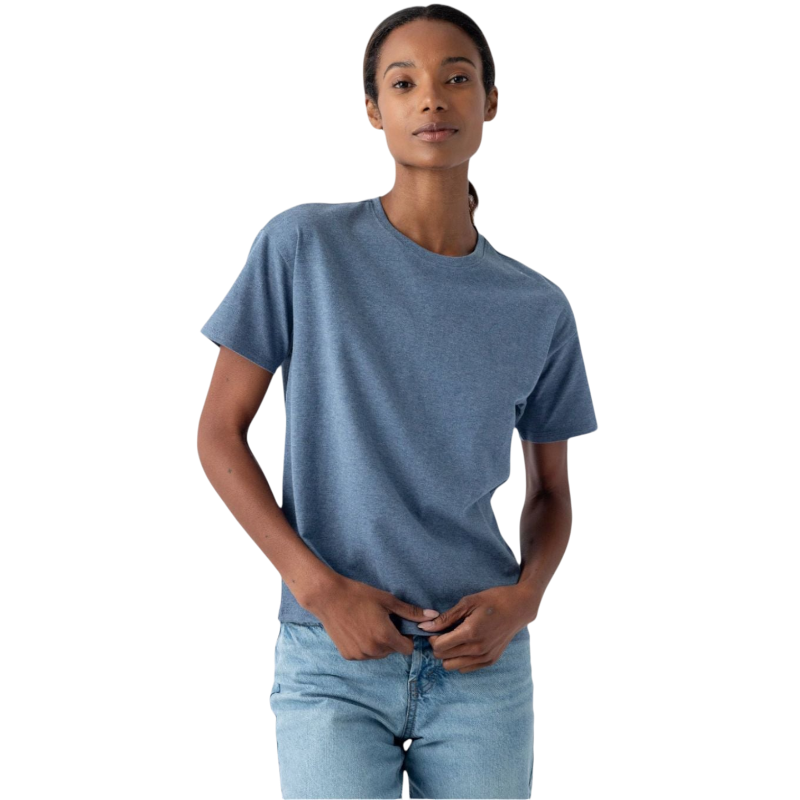 A person with long hair pulled back is wearing Cozy Earth's Women's All Day Tee, a blue short-sleeve t-shirt, along with light blue jeans. They are standing against a plain white background with a neutral expression, holding their hands near the waistband of their jeans. |Color:Deep Blue Heather