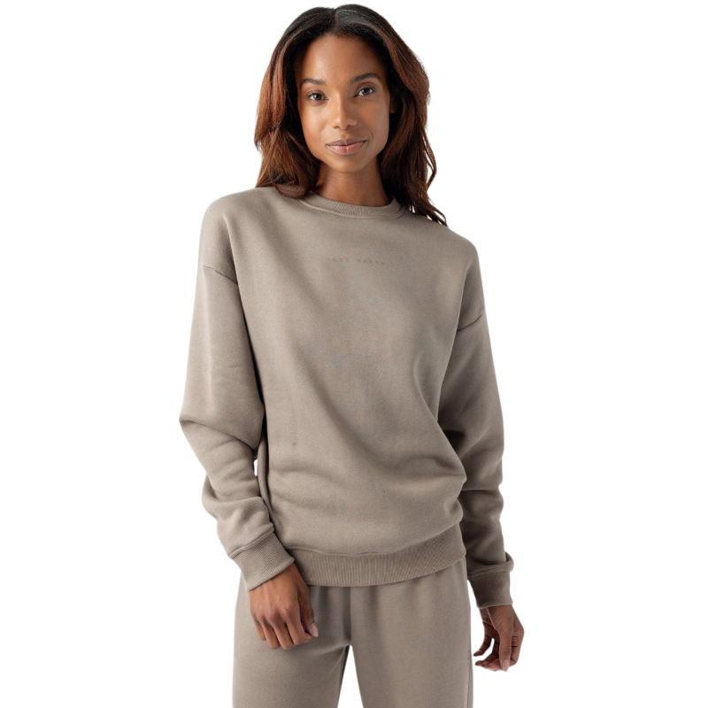 A person with long, wavy hair is wearing the Women's CityScape Crewneck and matching taupe-colored sweatpants from Cozy Earth. They are standing against a white background and smiling softly at the camera. The crewneck has subtle text on the chest. |Color:Ebony