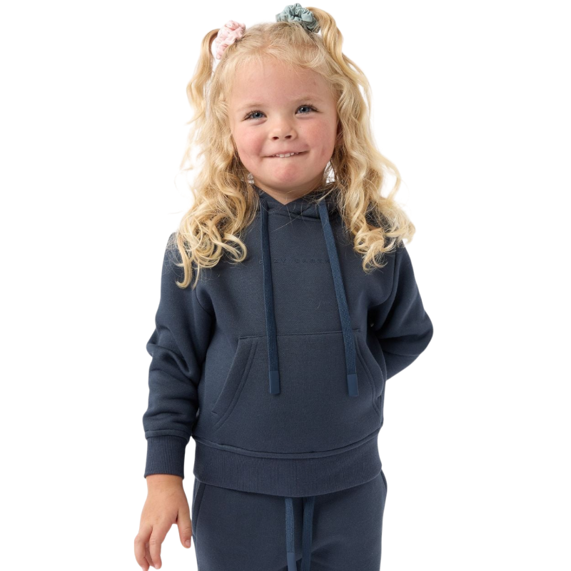 A young child with long blonde hair smiles while wearing a navy blue Kid's CityScape Hoodie by Cozy Earth and matching pants. She has two hair ties, one pink and one blue. The background is plain white. |Color:Eclipse