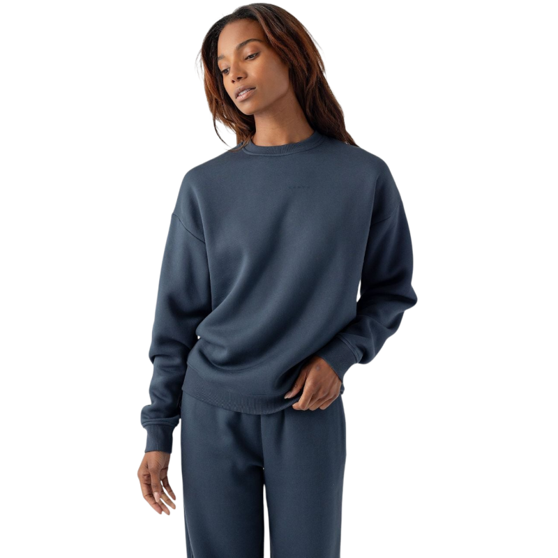 A person with long hair stands against a plain white background, dressed in Cozy Earth's dark blue Women's CityScape Crewneck sweatshirt and matching sweatpants. They have a relaxed expression with one hand resting casually by their side and the other touching the hem of the sweatshirt. |Color:Eclipse
