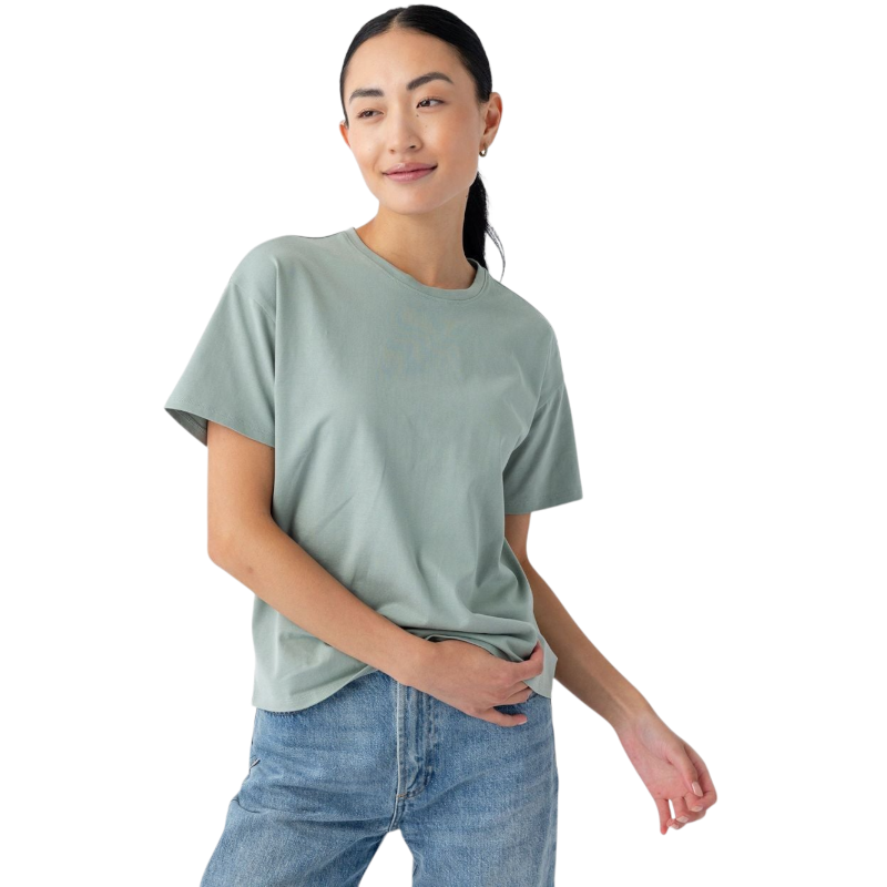 A person with long dark hair tied back is wearing the Women's All Day Tee in light green and blue jeans. They are standing against a plain white background, holding part of the t-shirt with one hand and smiling slightly. The t-shirt is by Cozy Earth. |Color:Haze