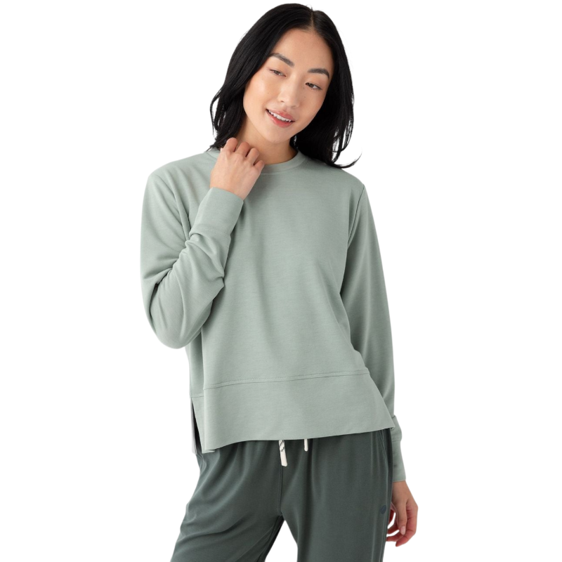 A person with long black hair is standing against a plain white background, wearing a Cozy Earth Women's StudioLite Crewneck in light green and dark green pants. They are looking down with a gentle smile, gently touching their neck with one hand. |Color:Haze
