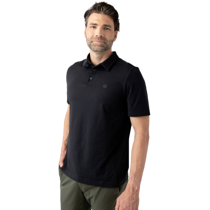 A man with short, dark hair and a beard wears a Men's Everyday Polo by Cozy Earth in black paired with dark green pants. He stands against a plain white background with his right hand in his pocket and a neutral expression on his face. |Color:Jet Black