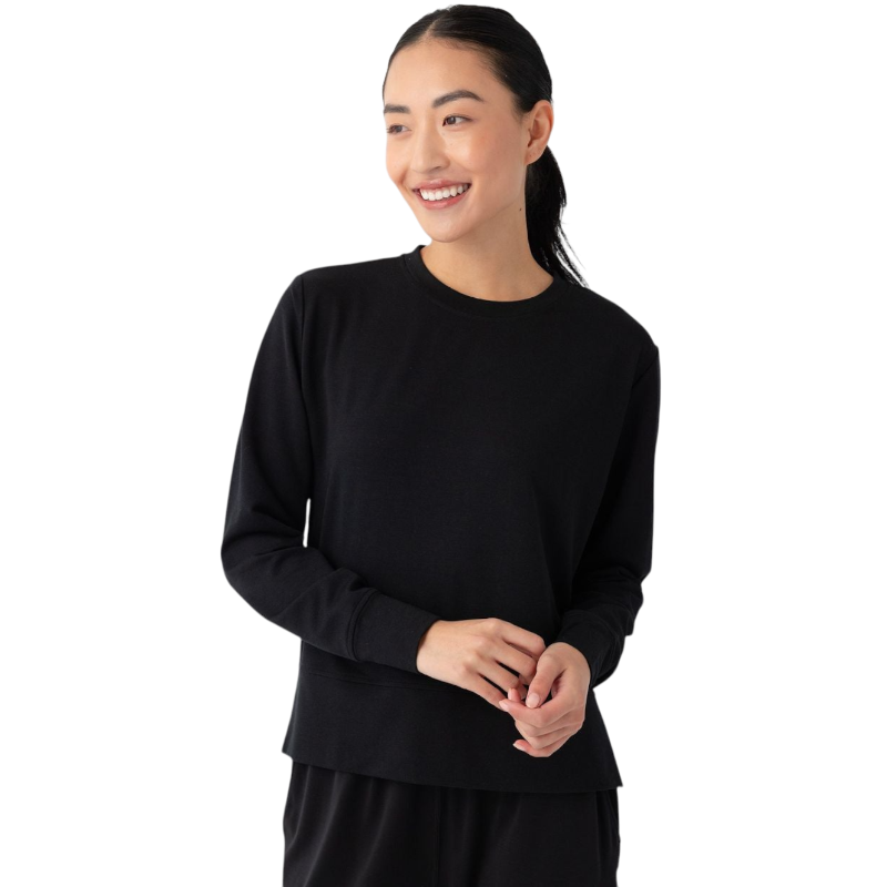 A person with long black hair is standing against a plain white background, wearing a Cozy Earth Women's StudioLite Crewneck in light green and dark green pants. They are looking down with a gentle smile, gently touching their neck with one hand. |Color:Haze