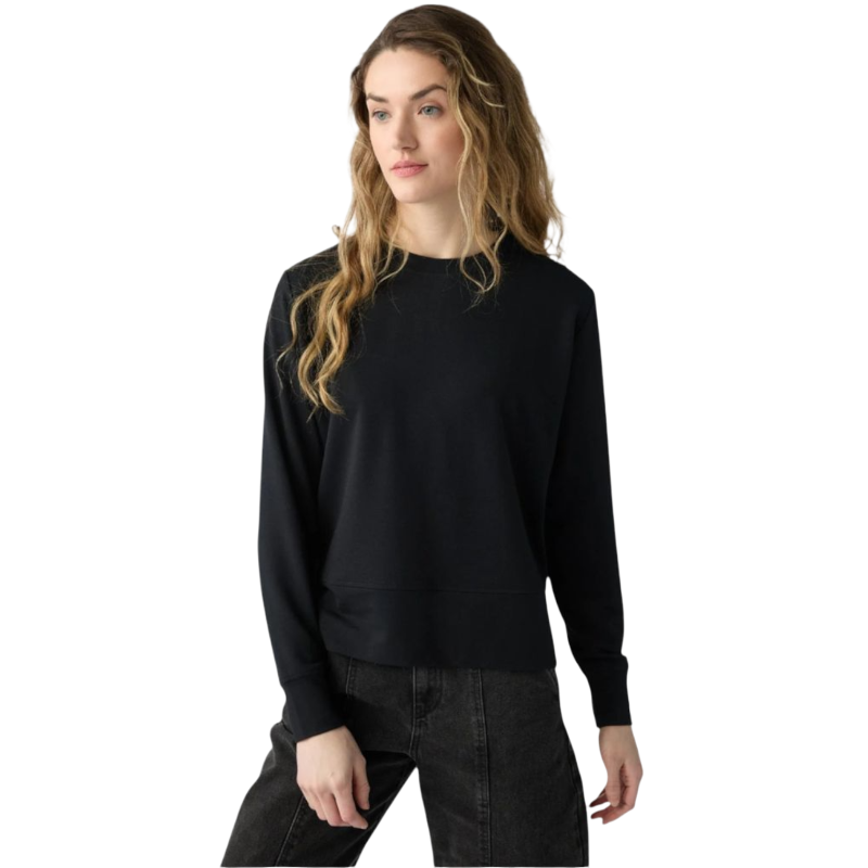 A woman with long wavy hair poses in a Cozy Earth Women's StudioLite Crewneck and dark jeans, with a neutral expression gazing slightly to the side against a simple white backdrop. |Color:Jet Black