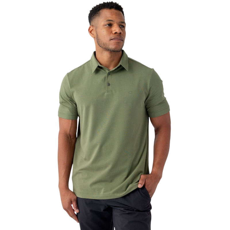 A person stands against a plain background wearing the Men's Everyday Polo by Cozy Earth in green, paired with black pants. They have short hair and are looking off to the side with one hand in their pocket. |Color:Juniper
