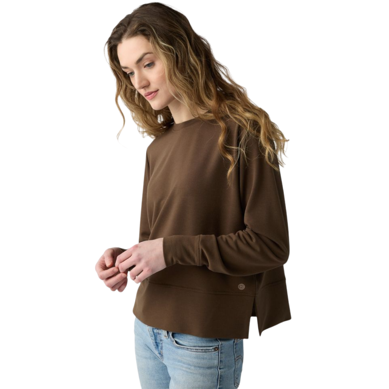 A person with long black hair is standing against a plain white background, wearing a Cozy Earth Women's StudioLite Crewneck in light green and dark green pants. They are looking down with a gentle smile, gently touching their neck with one hand. |Color:Haze