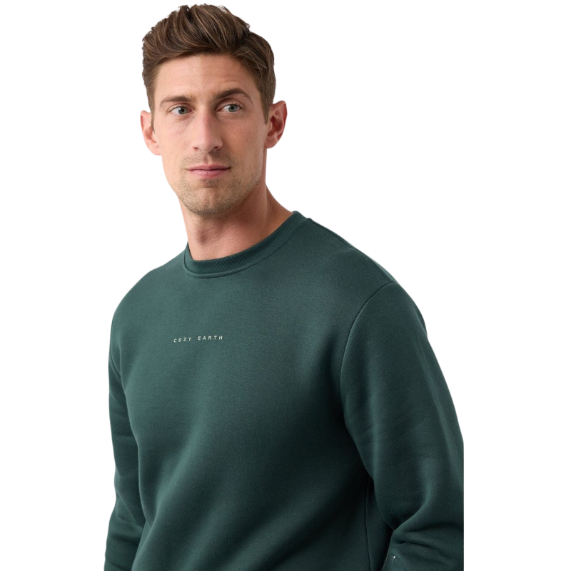 A man with short brown hair is wearing a dark green Men's CityScape Crewneck from Cozy Earth, featuring the words "COLD EARTH" printed on it. He is gazing slightly to the side against a plain white background. |Color:Spruce