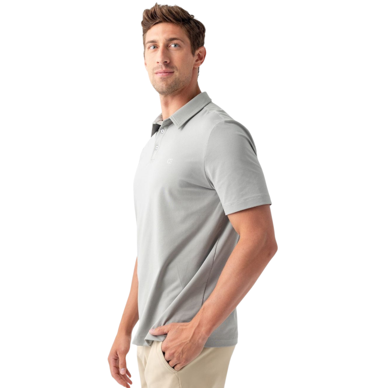 A person with short, light brown hair is wearing the Men's Everyday Polo by Cozy Earth in a light gray shade, paired with light beige pants. They are standing and slightly looking over their left shoulder with a neutral expression against a plain white background. |Color:Stone