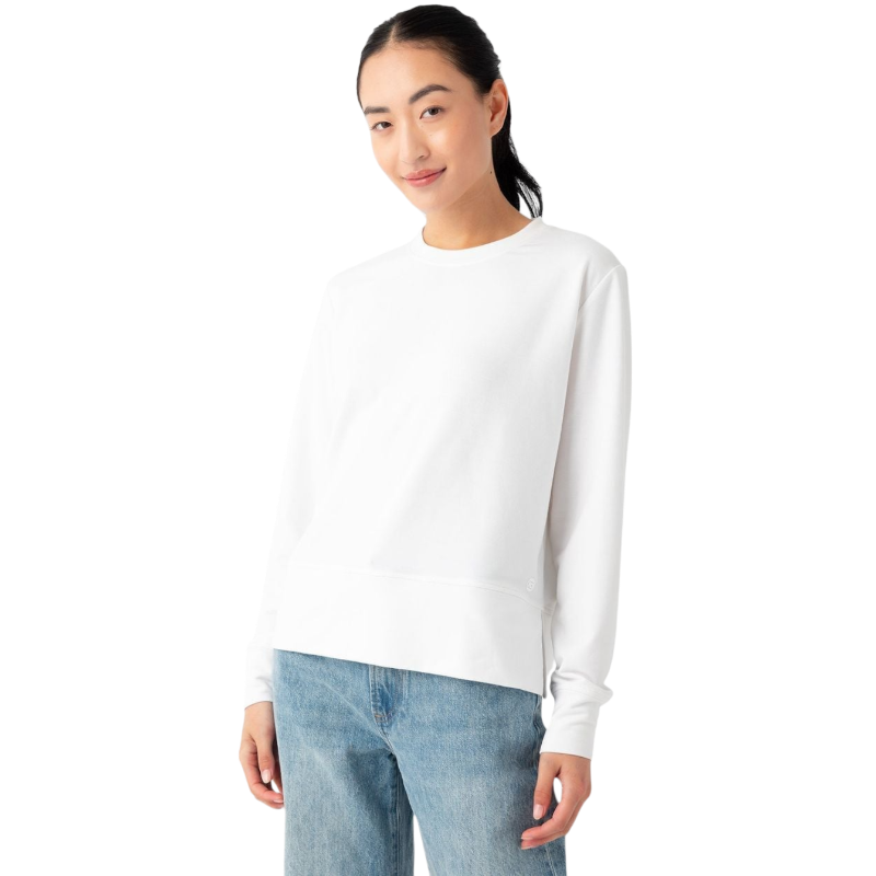 A person with long dark hair, wearing the Women's StudioLite Crewneck from Cozy Earth and light blue jeans, is standing against a plain white background. They are slightly smiling and have one hand in their pocket. |Color:White