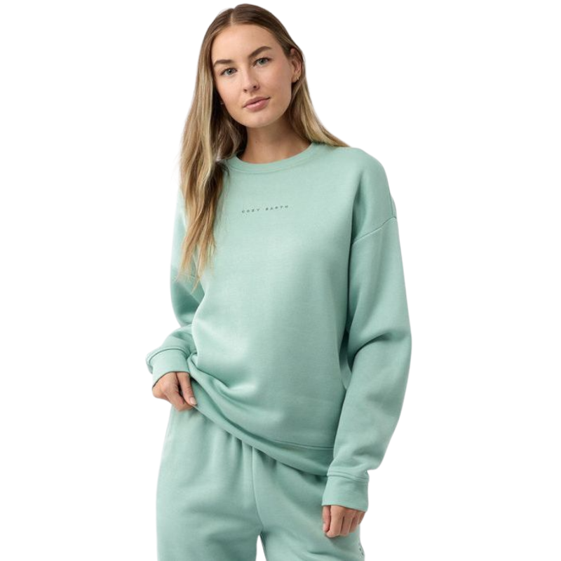 A person with long hair stands in a relaxed posture against a plain white background, wearing the Women's CityScape Crewneck from Cozy Earth in a matching teal set. The crewneck features text on it. |Color:Wintergreen