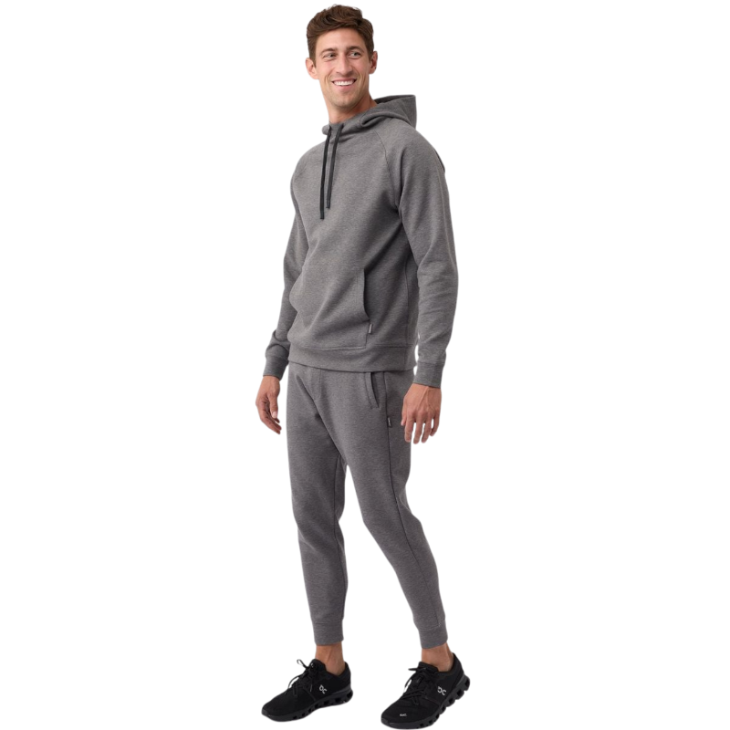 A person wearing the Men's StretchTech Hoodie from Cozy Earth, paired with matching gray pants, stands against a plain white background. The hoodie features drawstrings and a front pocket. The individual is looking forward with a neutral expression. |Color:Coal Heather