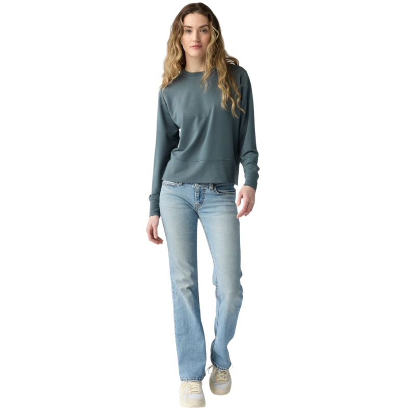 A person with long black hair is standing against a plain white background, wearing a Cozy Earth Women's StudioLite Crewneck in light green and dark green pants. They are looking down with a gentle smile, gently touching their neck with one hand. |Color:Haze