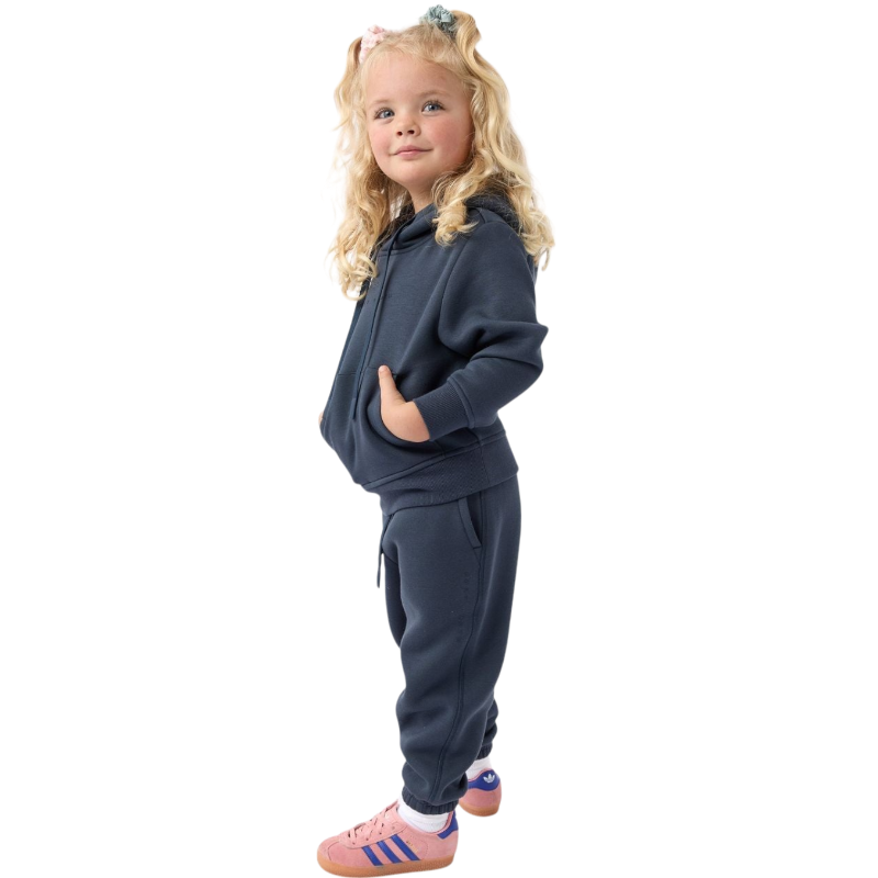 A young child with long blonde hair smiles while wearing a navy blue Kid's CityScape Hoodie by Cozy Earth and matching pants. She has two hair ties, one pink and one blue. The background is plain white. |Color:Eclipse