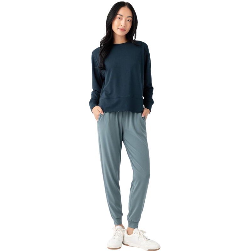 A person with long black hair is standing against a plain white background, wearing a Cozy Earth Women's StudioLite Crewneck in light green and dark green pants. They are looking down with a gentle smile, gently touching their neck with one hand. |Color:Haze
