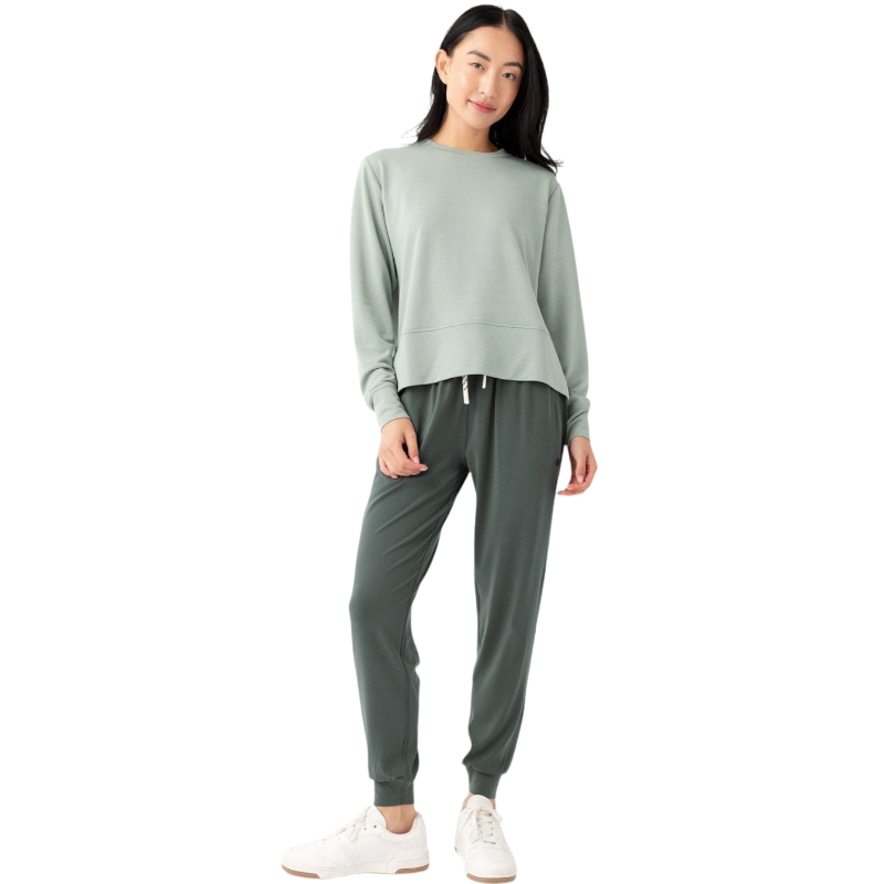 A person with long black hair is standing against a plain white background, wearing a Cozy Earth Women's StudioLite Crewneck in light green and dark green pants. They are looking down with a gentle smile, gently touching their neck with one hand. |Color:Haze