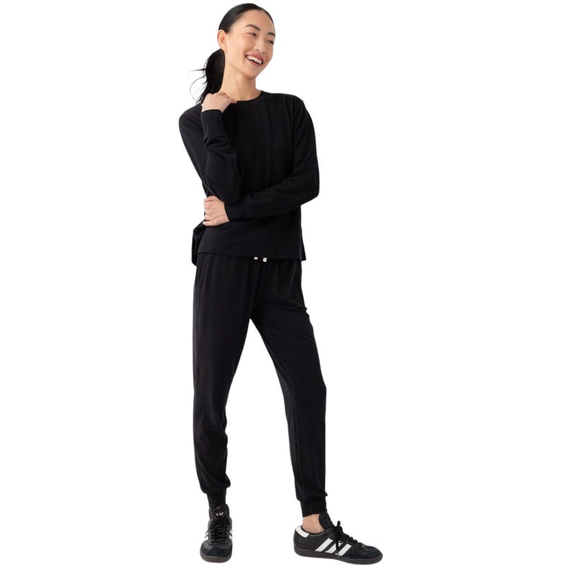 A person with long black hair is standing against a plain white background, wearing a Cozy Earth Women's StudioLite Crewneck in light green and dark green pants. They are looking down with a gentle smile, gently touching their neck with one hand. |Color:Haze