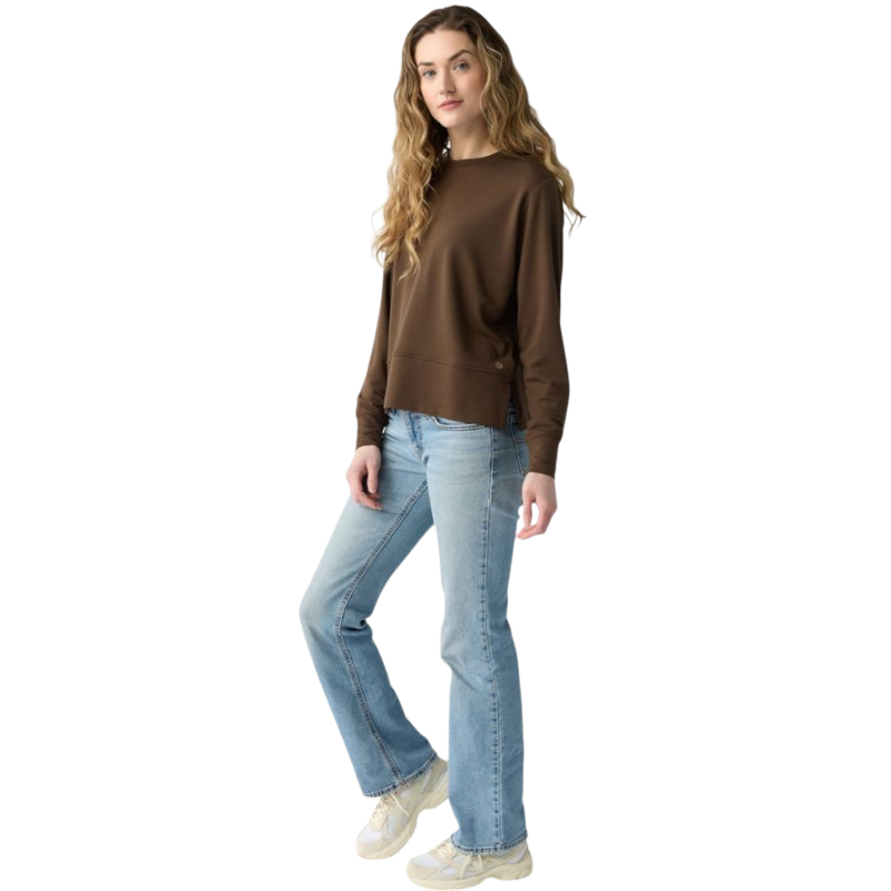 A person with long black hair is standing against a plain white background, wearing a Cozy Earth Women's StudioLite Crewneck in light green and dark green pants. They are looking down with a gentle smile, gently touching their neck with one hand. |Color:Haze