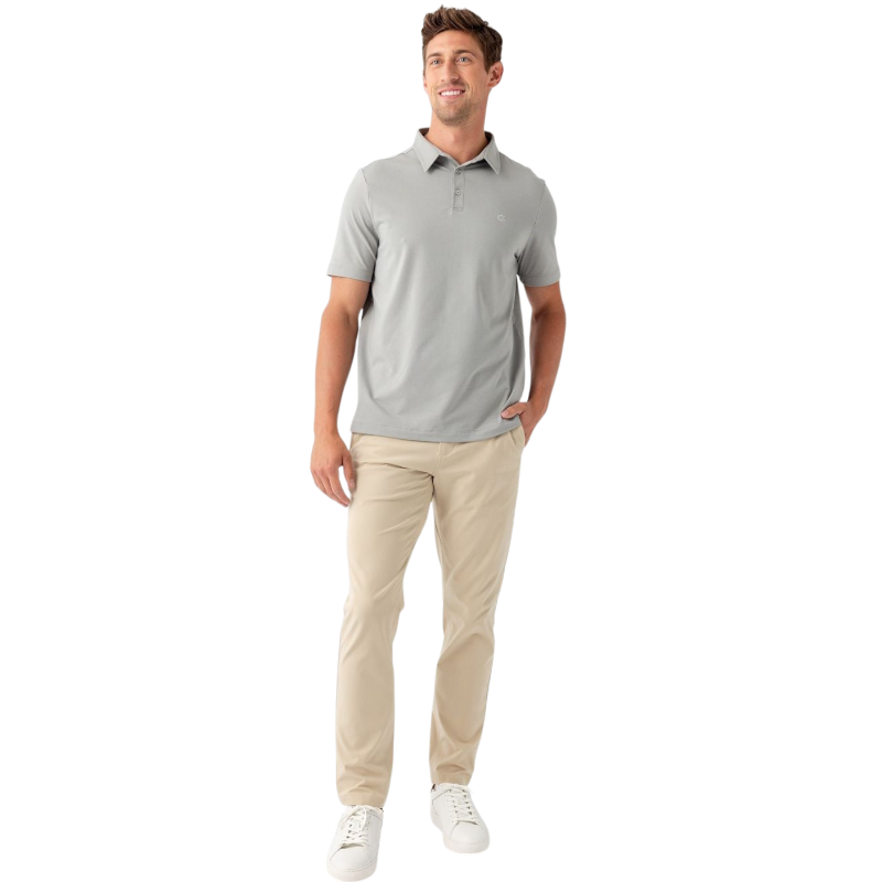 A person with short, light brown hair is wearing the Men's Everyday Polo by Cozy Earth in a light gray shade, paired with light beige pants. They are standing and slightly looking over their left shoulder with a neutral expression against a plain white background. |Color:Stone