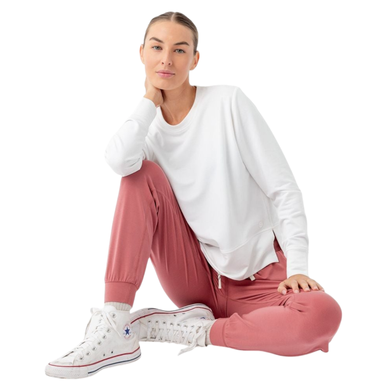 A person with long black hair is standing against a plain white background, wearing a Cozy Earth Women's StudioLite Crewneck in light green and dark green pants. They are looking down with a gentle smile, gently touching their neck with one hand. |Color:Haze