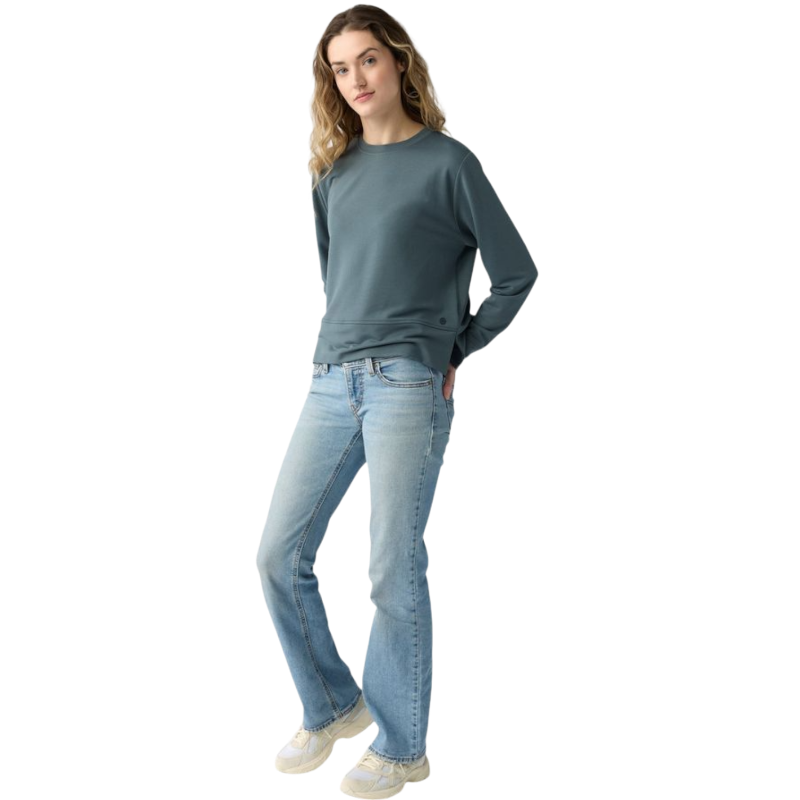 A person with long black hair is standing against a plain white background, wearing a Cozy Earth Women's StudioLite Crewneck in light green and dark green pants. They are looking down with a gentle smile, gently touching their neck with one hand. |Color:Haze