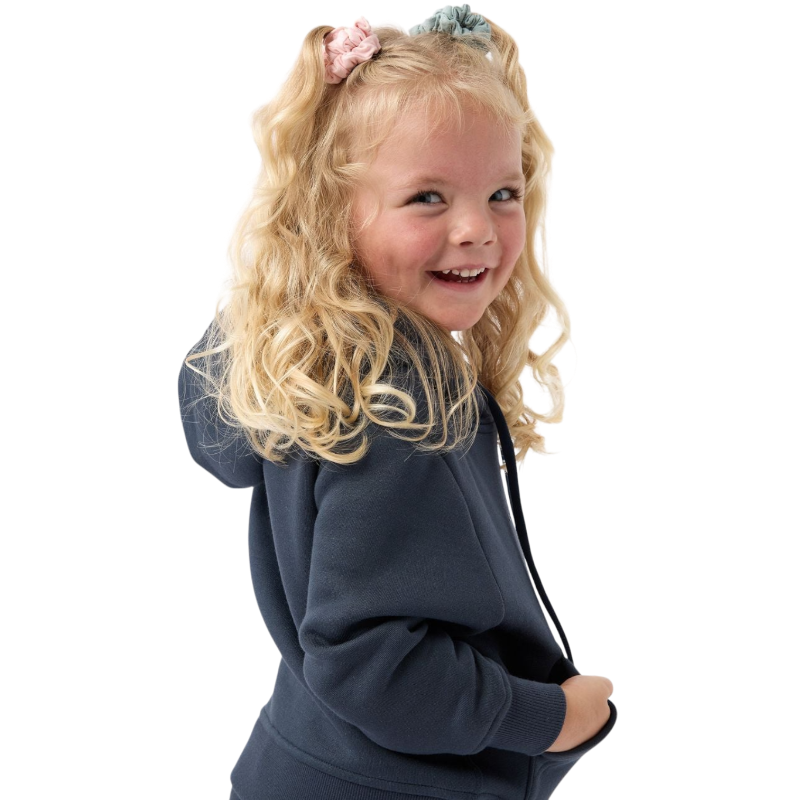 A young child with long blonde hair smiles while wearing a navy blue Kid's CityScape Hoodie by Cozy Earth and matching pants. She has two hair ties, one pink and one blue. The background is plain white. |Color:Eclipse