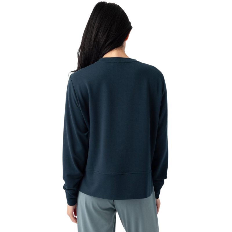 A person with long black hair is standing against a plain white background, wearing a Cozy Earth Women's StudioLite Crewneck in light green and dark green pants. They are looking down with a gentle smile, gently touching their neck with one hand. |Color:Haze