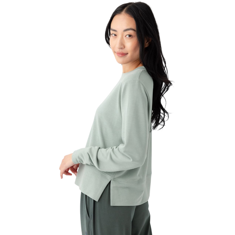 A person with long black hair is standing against a plain white background, wearing a Cozy Earth Women's StudioLite Crewneck in light green and dark green pants. They are looking down with a gentle smile, gently touching their neck with one hand. |Color:Haze