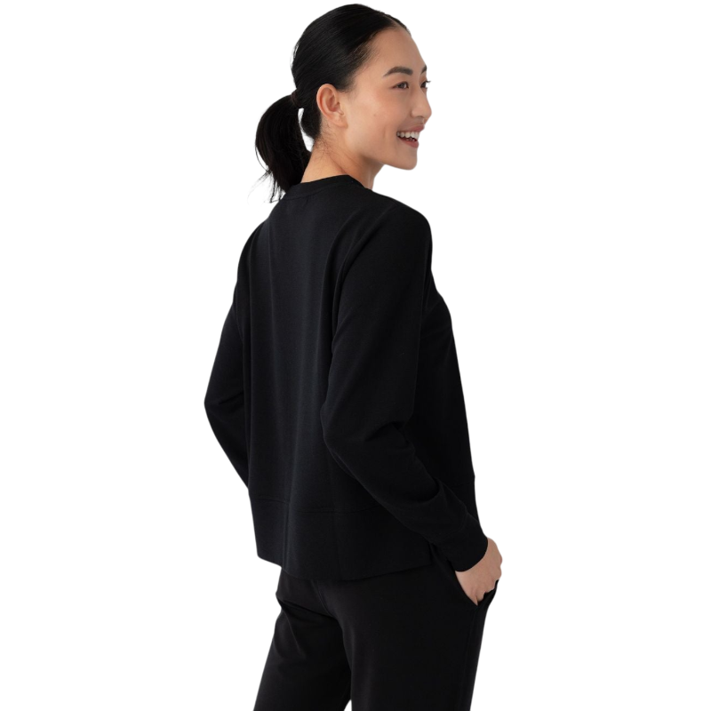 A person with long black hair is standing against a plain white background, wearing a Cozy Earth Women's StudioLite Crewneck in light green and dark green pants. They are looking down with a gentle smile, gently touching their neck with one hand. |Color:Haze