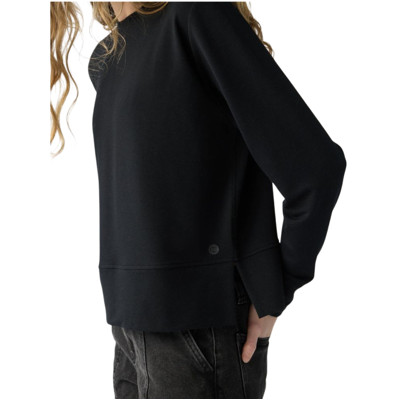 A person with long black hair is standing against a plain white background, wearing a Cozy Earth Women's StudioLite Crewneck in light green and dark green pants. They are looking down with a gentle smile, gently touching their neck with one hand. |Color:Haze