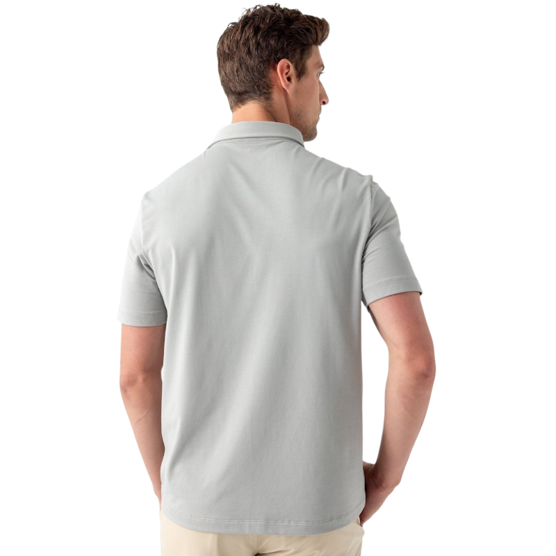 A person with short, light brown hair is wearing the Men's Everyday Polo by Cozy Earth in a light gray shade, paired with light beige pants. They are standing and slightly looking over their left shoulder with a neutral expression against a plain white background. |Color:Stone