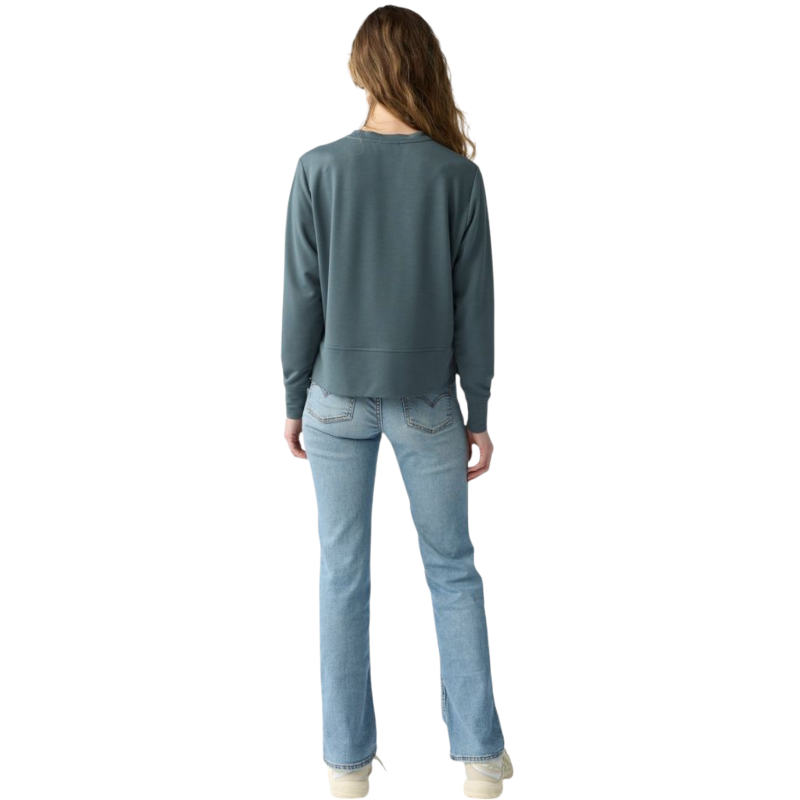 A person with long black hair is standing against a plain white background, wearing a Cozy Earth Women's StudioLite Crewneck in light green and dark green pants. They are looking down with a gentle smile, gently touching their neck with one hand. |Color:Haze