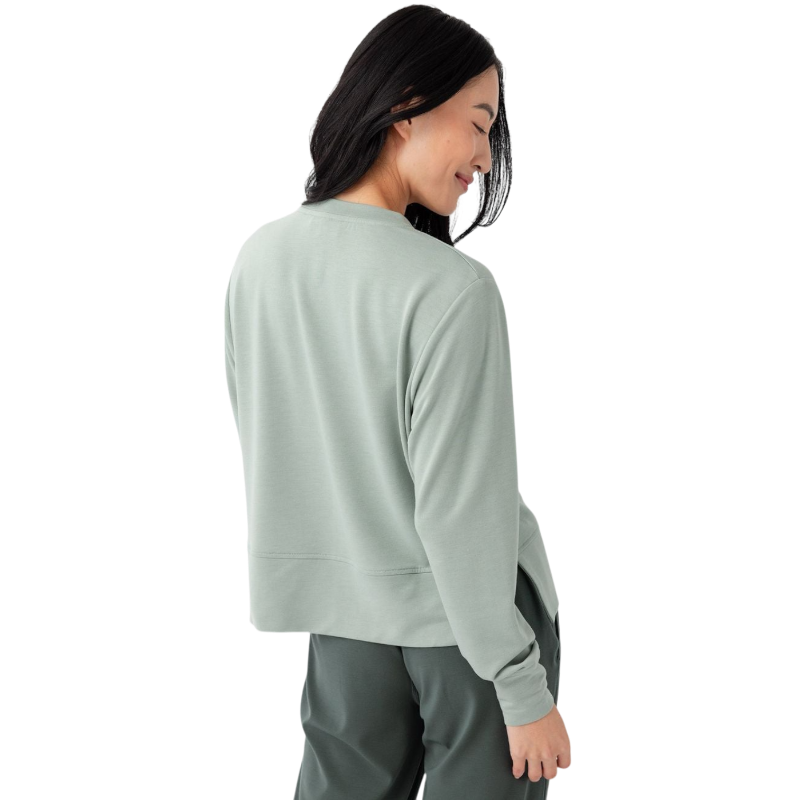 A person with long black hair is standing against a plain white background, wearing a Cozy Earth Women's StudioLite Crewneck in light green and dark green pants. They are looking down with a gentle smile, gently touching their neck with one hand. |Color:Haze