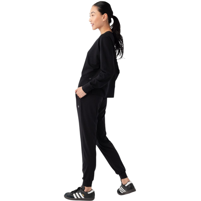 A person with long black hair is standing against a plain white background, wearing a Cozy Earth Women's StudioLite Crewneck in light green and dark green pants. They are looking down with a gentle smile, gently touching their neck with one hand. |Color:Haze