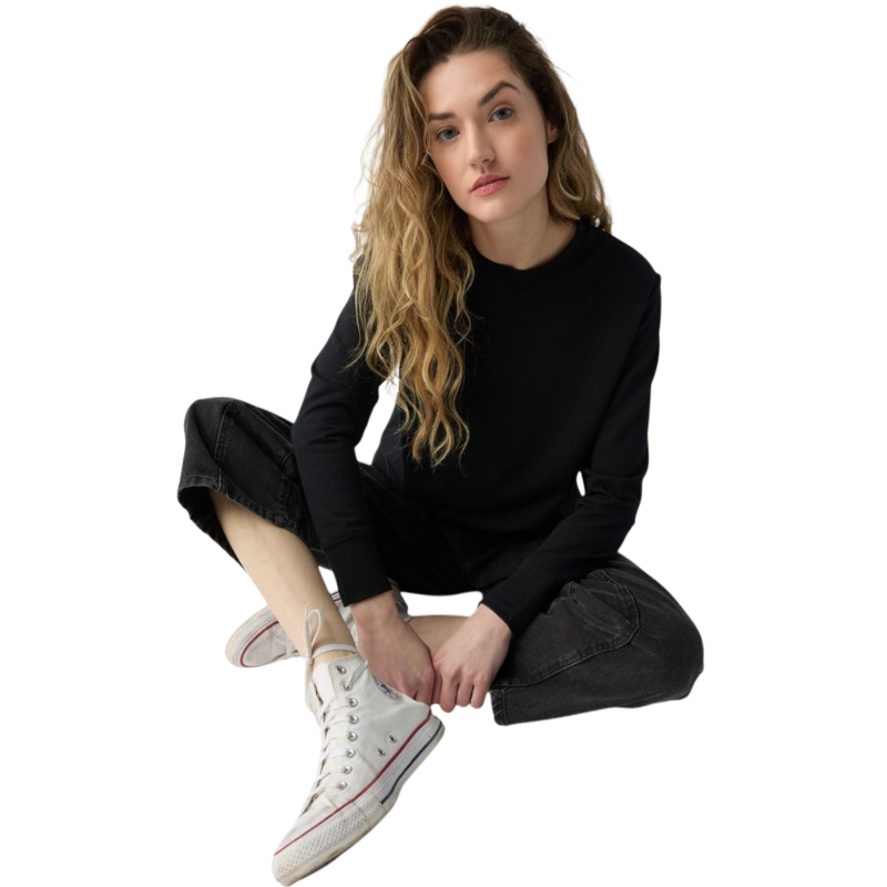 A person with long black hair is standing against a plain white background, wearing a Cozy Earth Women's StudioLite Crewneck in light green and dark green pants. They are looking down with a gentle smile, gently touching their neck with one hand. |Color:Haze