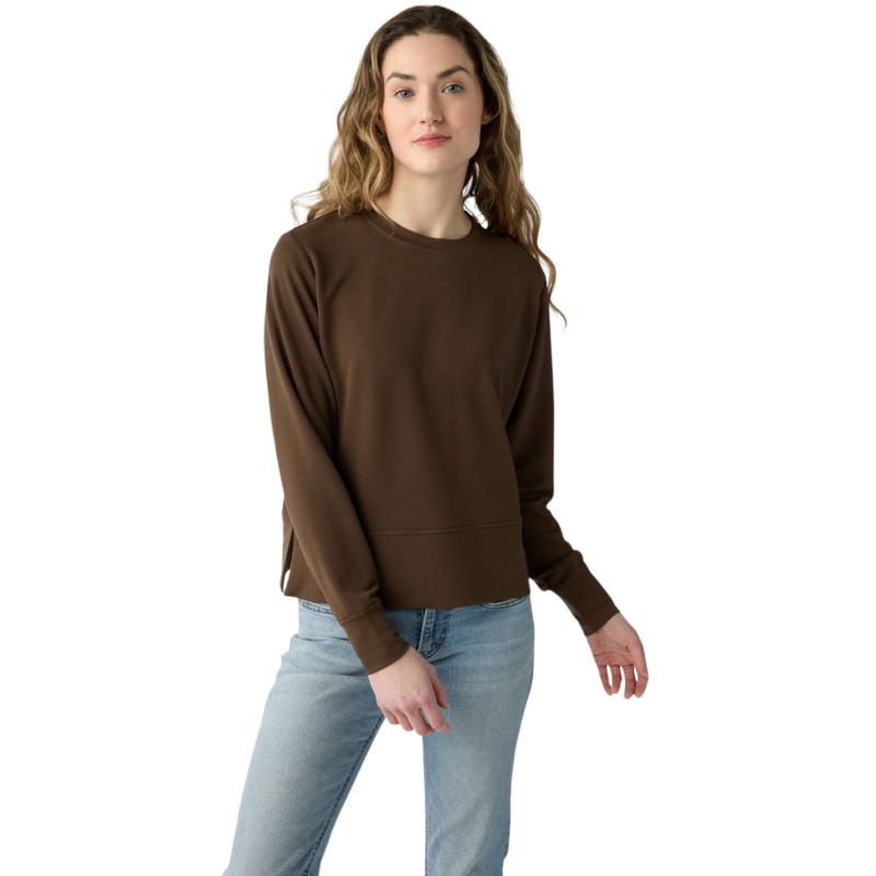 A person with long black hair is standing against a plain white background, wearing a Cozy Earth Women's StudioLite Crewneck in light green and dark green pants. They are looking down with a gentle smile, gently touching their neck with one hand. |Color:Haze