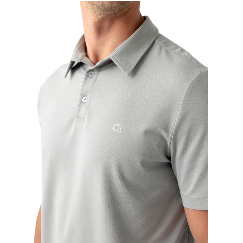 A person with short, light brown hair is wearing the Men's Everyday Polo by Cozy Earth in a light gray shade, paired with light beige pants. They are standing and slightly looking over their left shoulder with a neutral expression against a plain white background. |Color:Stone