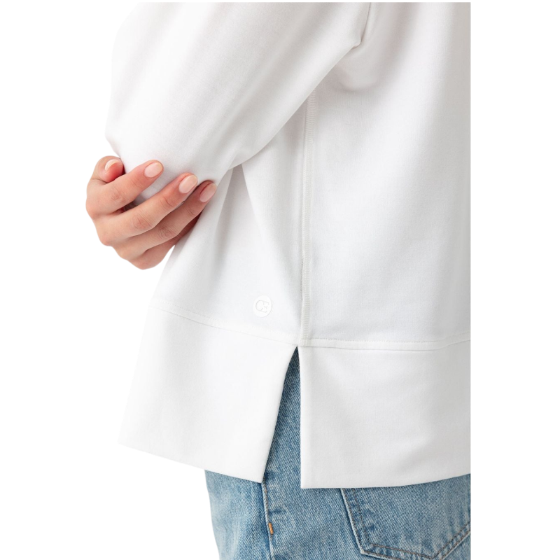 A person with long black hair is standing against a plain white background, wearing a Cozy Earth Women's StudioLite Crewneck in light green and dark green pants. They are looking down with a gentle smile, gently touching their neck with one hand. |Color:Haze