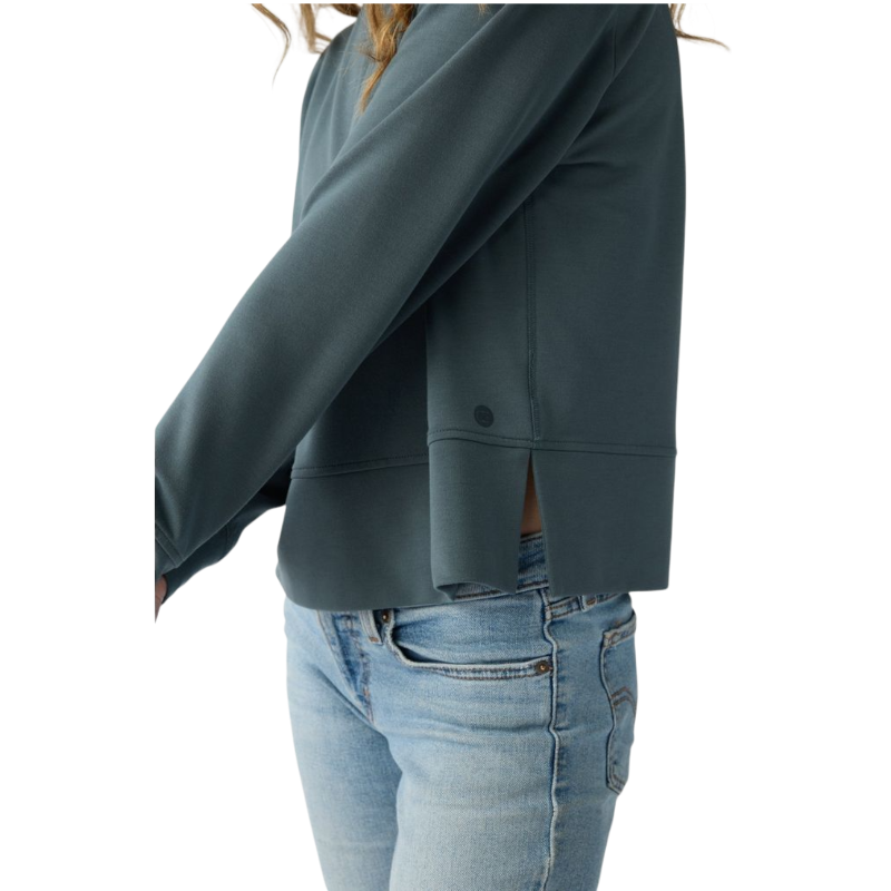 A person with long black hair is standing against a plain white background, wearing a Cozy Earth Women's StudioLite Crewneck in light green and dark green pants. They are looking down with a gentle smile, gently touching their neck with one hand. |Color:Haze