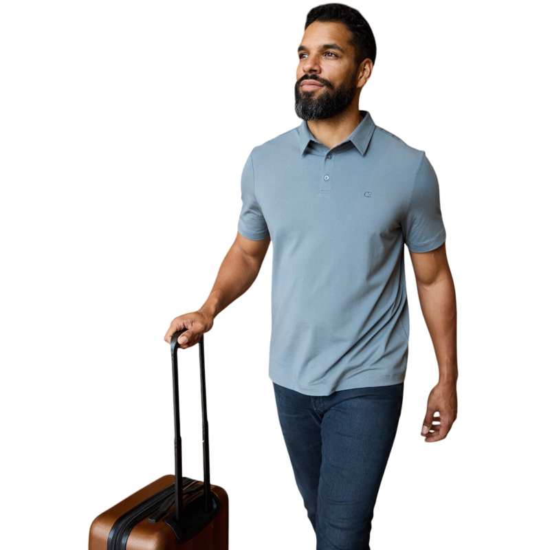 A person with short, light brown hair is wearing the Men's Everyday Polo by Cozy Earth in a light gray shade, paired with light beige pants. They are standing and slightly looking over their left shoulder with a neutral expression against a plain white background. |Color:Stone