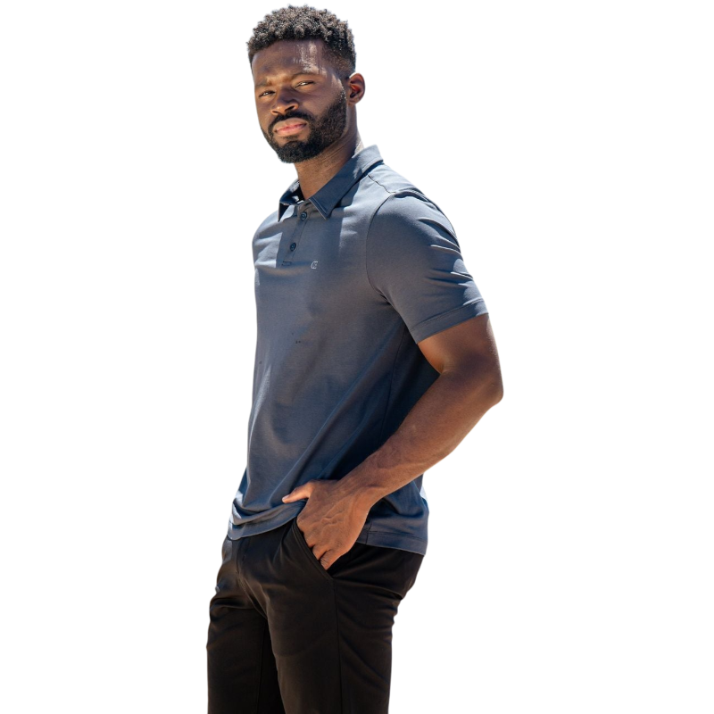 A person with short, light brown hair is wearing the Men's Everyday Polo by Cozy Earth in a light gray shade, paired with light beige pants. They are standing and slightly looking over their left shoulder with a neutral expression against a plain white background. |Color:Stone