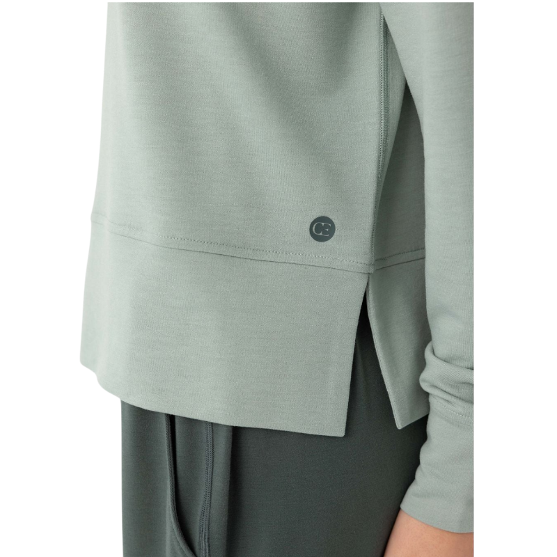 A person with long black hair is standing against a plain white background, wearing a Cozy Earth Women's StudioLite Crewneck in light green and dark green pants. They are looking down with a gentle smile, gently touching their neck with one hand. |Color:Haze