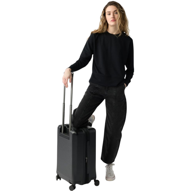 A person with long black hair is standing against a plain white background, wearing a Cozy Earth Women's StudioLite Crewneck in light green and dark green pants. They are looking down with a gentle smile, gently touching their neck with one hand. |Color:Haze