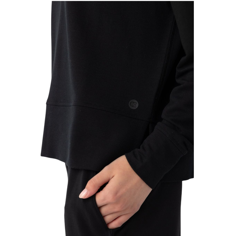 A person with long black hair is standing against a plain white background, wearing a Cozy Earth Women's StudioLite Crewneck in light green and dark green pants. They are looking down with a gentle smile, gently touching their neck with one hand. |Color:Haze