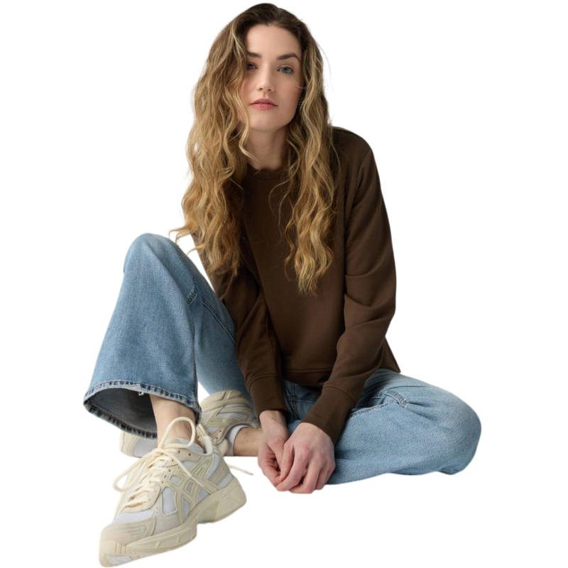 A person with long black hair is standing against a plain white background, wearing a Cozy Earth Women's StudioLite Crewneck in light green and dark green pants. They are looking down with a gentle smile, gently touching their neck with one hand. |Color:Haze