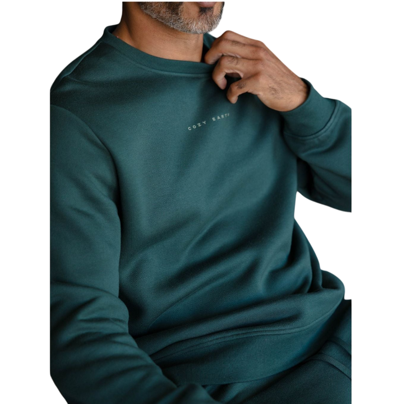 A man with short brown hair is wearing a dark green Men's CityScape Crewneck from Cozy Earth, featuring the words "COLD EARTH" printed on it. He is gazing slightly to the side against a plain white background. |Color:Spruce